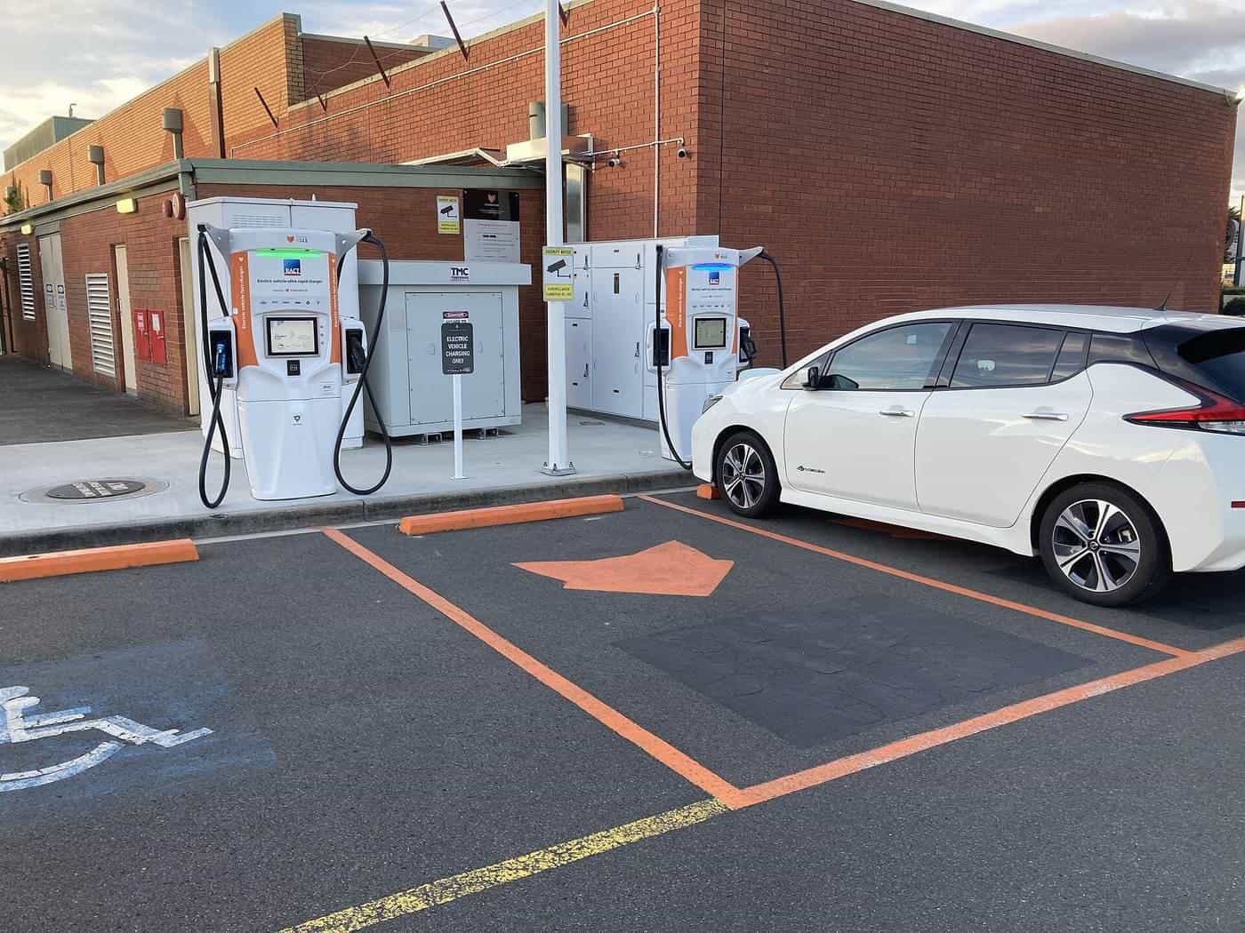 A white electric hatchback parked at a two-bay fast-charging station with orange markings in Kings Meadows, Tasmania
